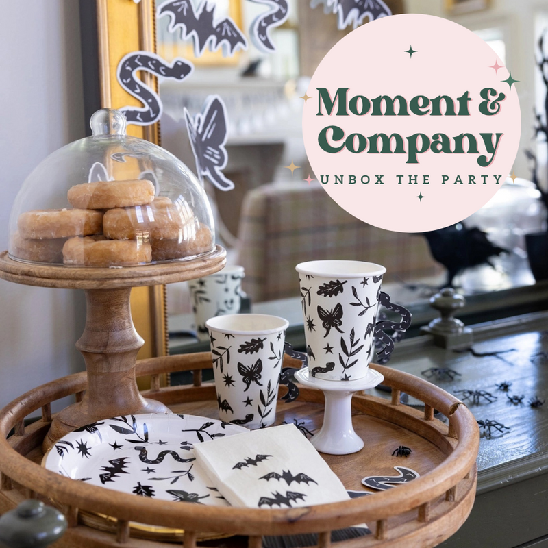 A stylish Halloween party setup featuring black and white themed plates, cups, and napkins with spooky designs, displayed alongside donuts on a wooden tray.