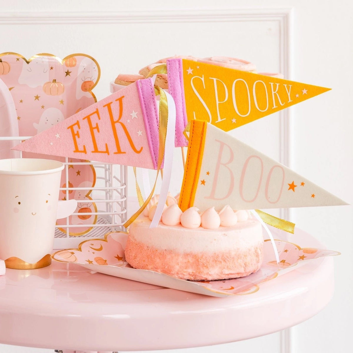 Close-up of Halloween party table featuring ghost plates, pink ghost cups, and mini felt pennants reading Spooky, Eek, and Boo ideal for a Halloween boo basket.