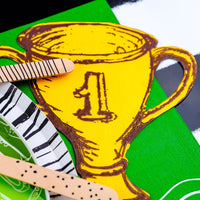 Golden trophy-shaped paper napkin alongside green and white sports-themed plates and wooden cutlery, a great addition to any sports themed birthday party.