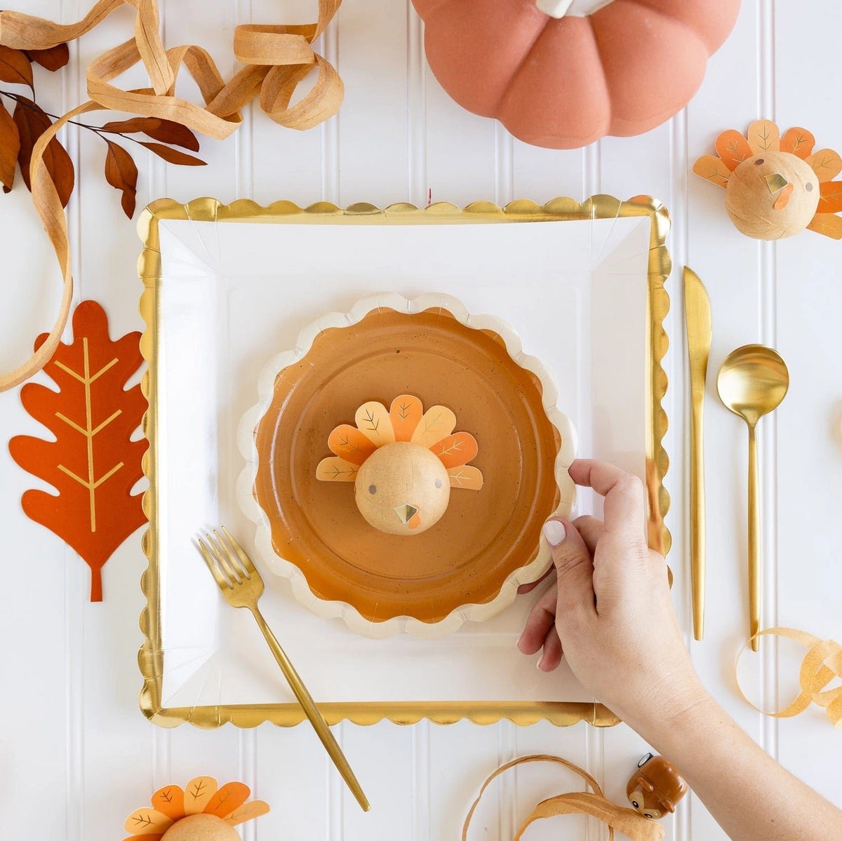 A beautifully set Thanksgiving table featuring a gold scalloped-edge paper plate, a turkey-themed place setting, and festive fall decorations. Perfect for adding a touch of elegance to your Thanksgiving feast.