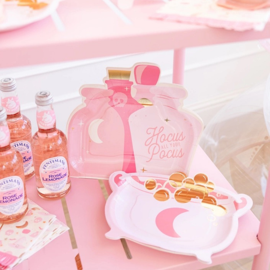 Close-up of pink potion bottle and cauldron-shaped Halloween paper plates on a pink table, perfect for Hocus Pocus decor and pink Halloween decor.