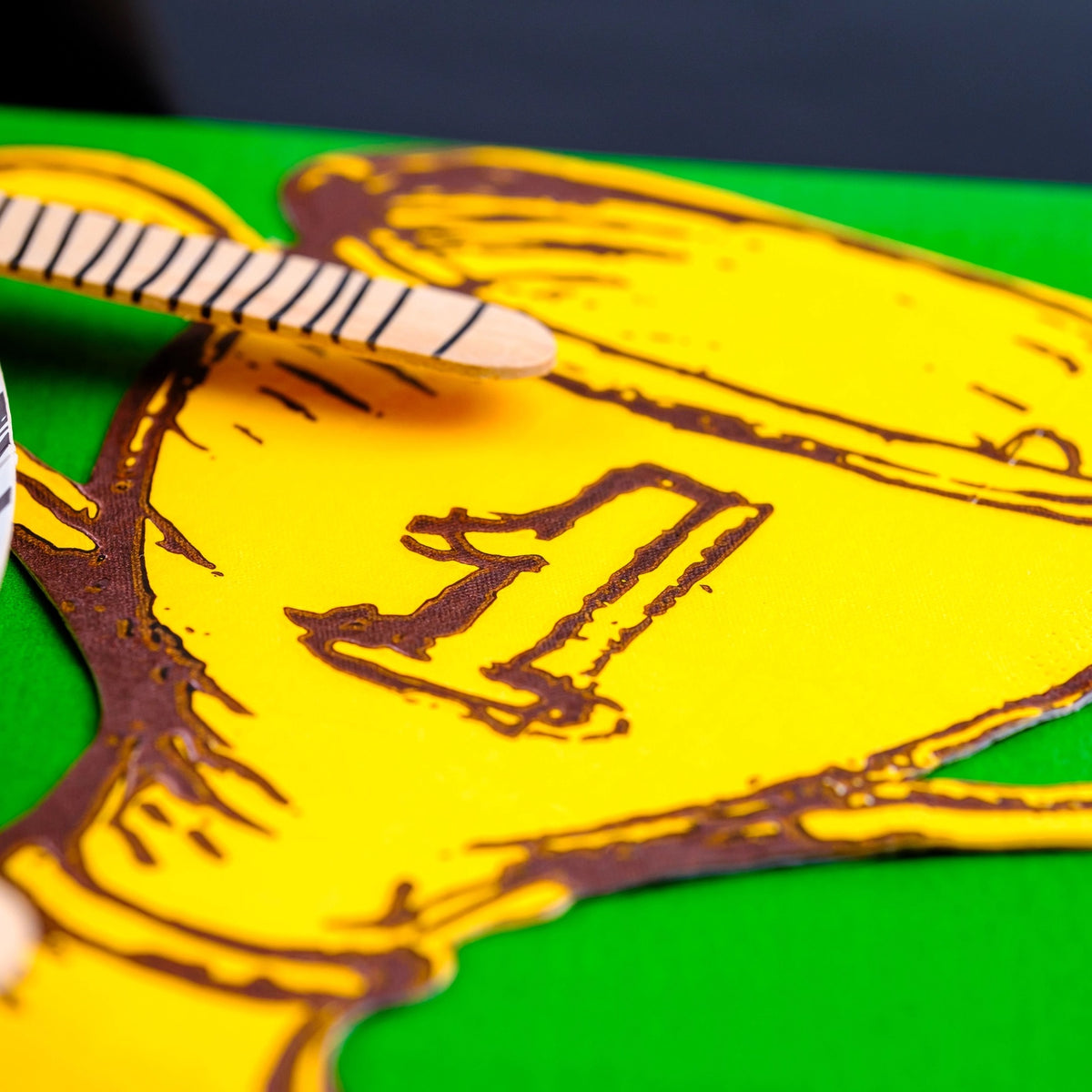 Detailed view of a golden trophy-shaped paper napkin on a green surface with striped plates and wooden cutlery, perfect for a sports themed birthday party.