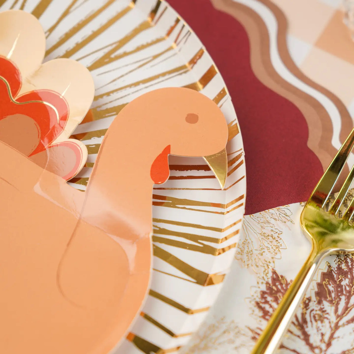 A festive Thanksgiving tablescape featuring a maroon charger plate, a gold-accented dinner plate, and a paper plate turkey on top, ready for desserts.