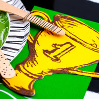 Close-up of a golden trophy-shaped paper napkin placed on a green background with wooden cutlery, ideal for a sports themed birthday party.