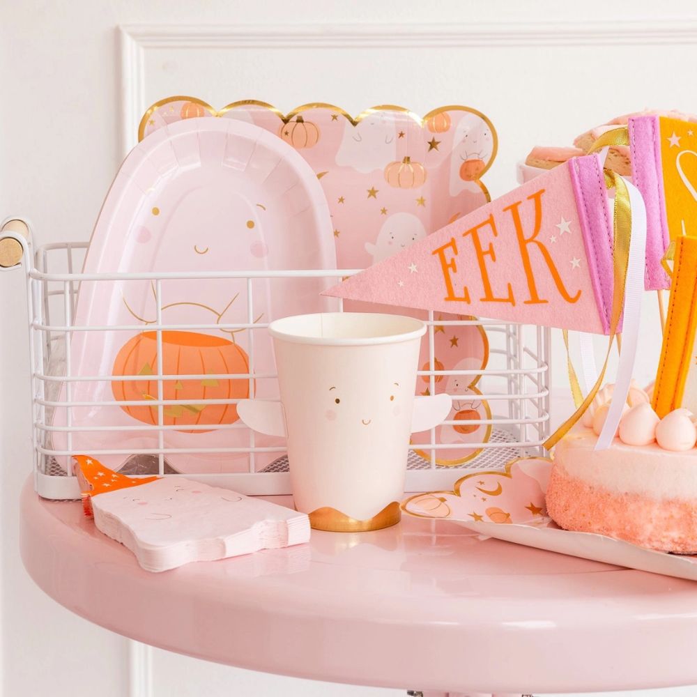 Close-up of ghost-shaped paper plates, a pink ghost cup, and Halloween-themed napkins arranged on a pastel pink table