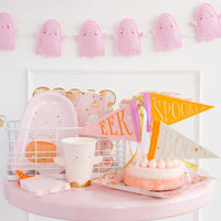 A display of cute Halloween party supplies featuring ghost-shaped paper plates, pink ghost cups, and mini pennants with EEK, SPOOKY, and BOO in vibrant colors.