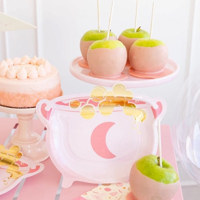 Pink cauldron-shaped Halloween paper plate with gold potion bubbles on a dessert table with pink caramel apples and cake.