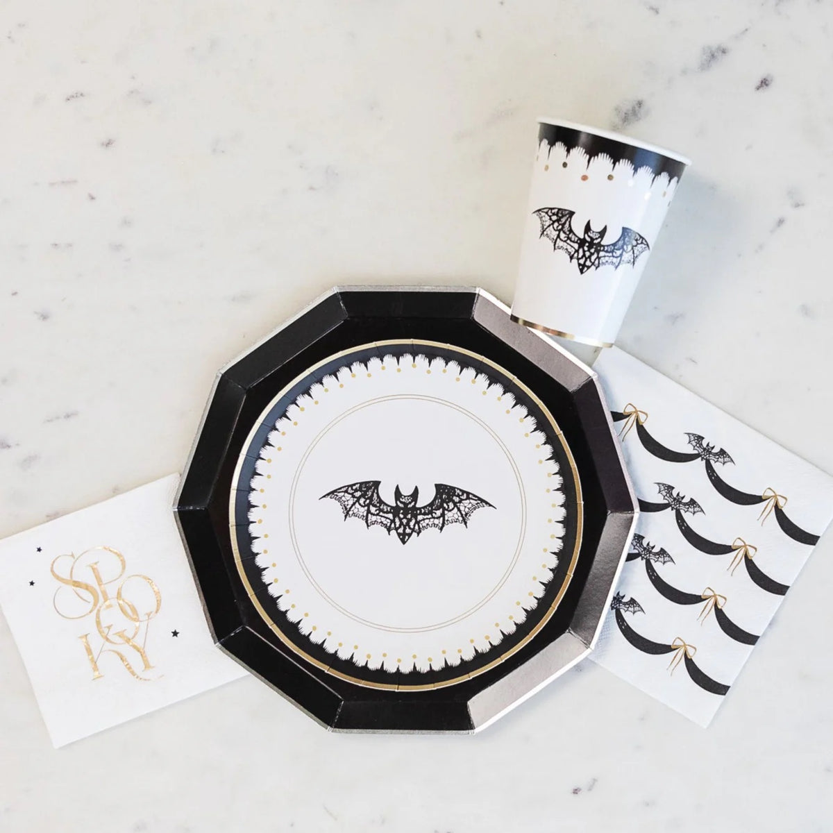 A flat lay of elegant Halloween decor featuring a black and white Halloween napkin with intricate bat designs and gold bows. The matching Halloween plate and cup complete the stylish look for a spooky, refined celebration.