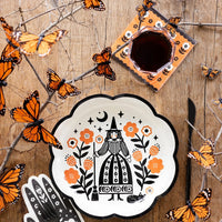 Table setting featuring Halloween paper cocktail napkins with orange and black floral design, matching plate with a witch illustration, surrounded by orange butterflies.