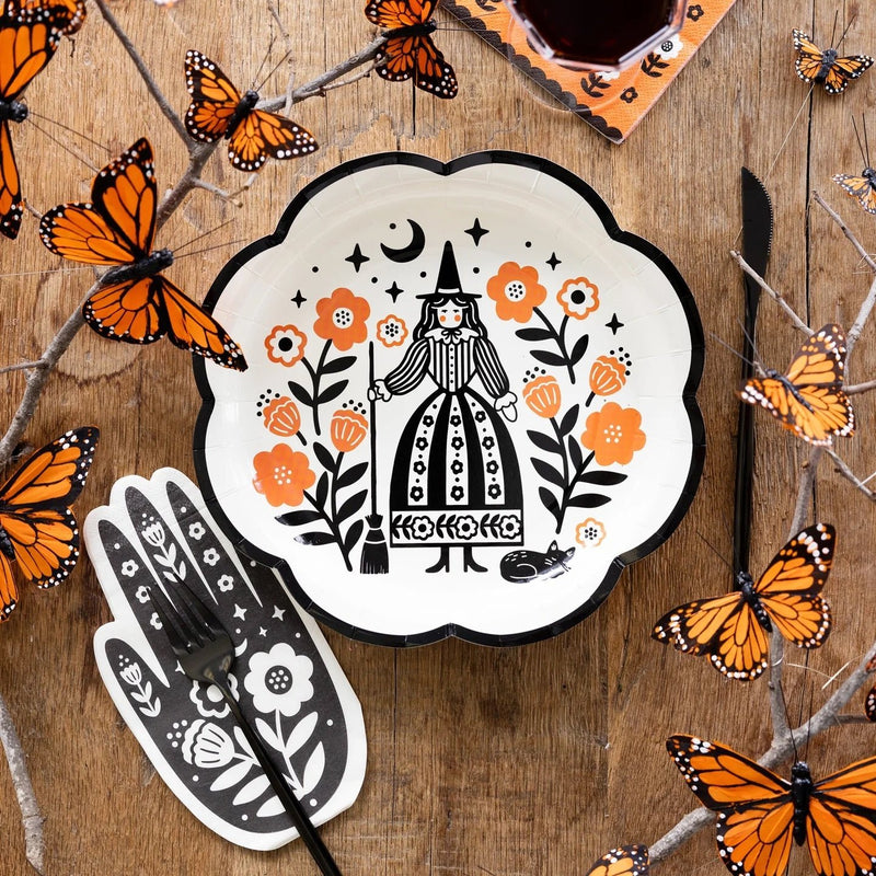 Table setting featuring a black and white witch-themed plate, a hand-shaped Halloween paper napkin, and orange paper napkins with floral designs, all surrounded by orange butterflies.