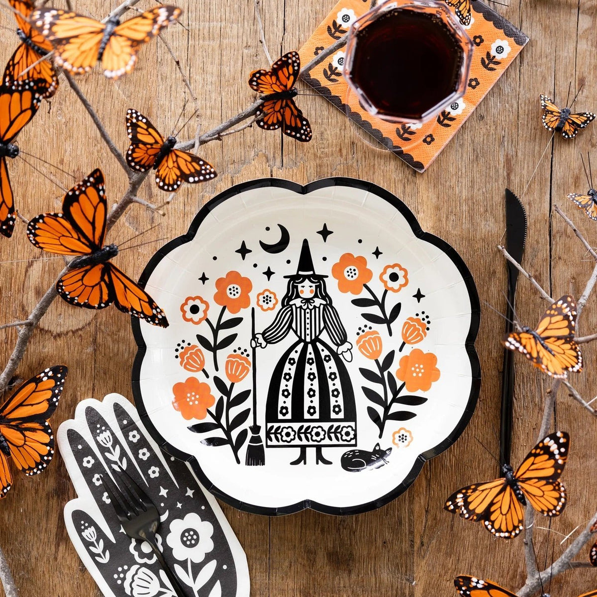 Halloween table setup featuring a witch-themed plate, hand-shaped napkin, and floral orange napkins, with orange butterflies decorating the scene.