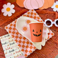 A Halloween table setup featuring an orange jack-o'-lantern paper cup, paired with checkered plates and 'Trick or Treat' napkins for a fun and spooky party.