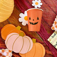 A Halloween-themed table display with a cheerful orange jack-o'-lantern paper cup as the centerpiece, surrounded by pumpkin-shaped napkins and Halloween decor.