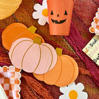 A Halloween table setting featuring orange and pink pumpkin-shaped paper napkins, paired with Halloween decor including a jack-o'-lantern cup and checkered plates.