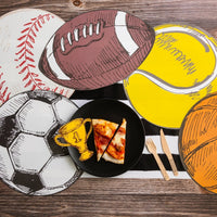 Collection of sports-themed placemats including a football, soccer ball, baseball, tennis ball, and basketball, arranged on a table with pizza and party decorations. Football placemat stands out as part of the football party decor.