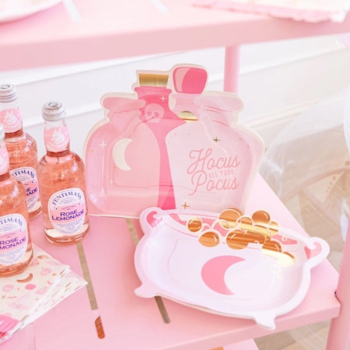 Pink potion bottle and cauldron-shaped Halloween paper plates on a pink table with rose lemonade bottles.