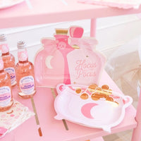 Pink potion bottle and cauldron-shaped Halloween paper plates on a pink table with rose lemonade bottles.