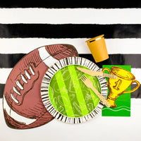 Football paper plate set up with a black and white striped table runner, golden trophy napkin, and wooden cutlery, ideal decorations for a tailgate party.