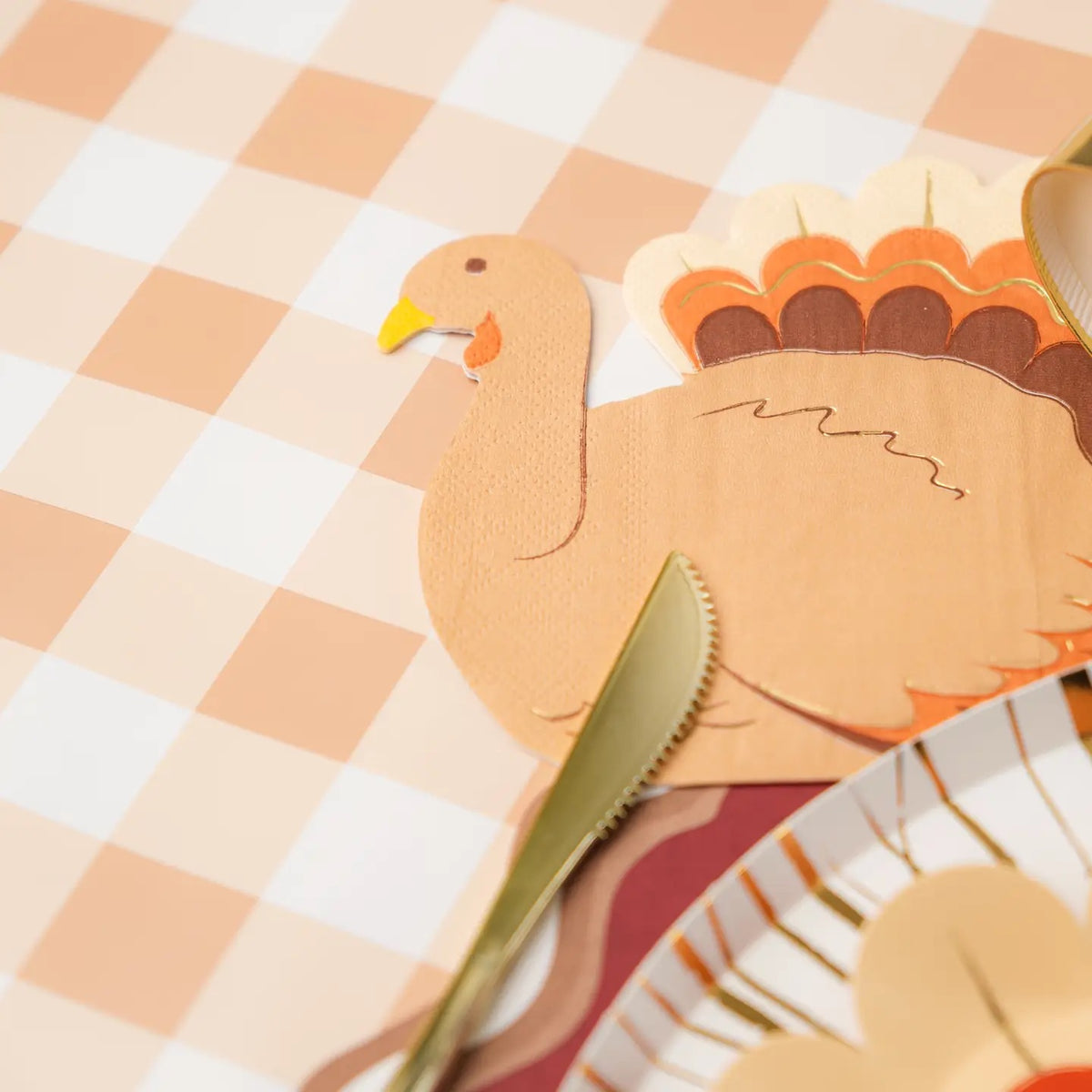  A close-up of a turkey napkin placed next to gold cutlery on a gingham tablecloth. These Thanksgiving paper napkins are the perfect decorative and functional addition to your holiday table.