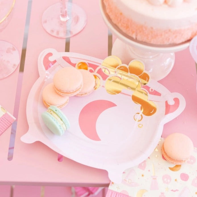 Pink cauldron-shaped Halloween paper plate with gold potion bubbles holding macarons on a pink dessert table.