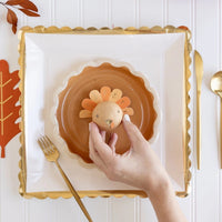 A close-up of a Thanksgiving place setting featuring a gold scalloped-edge paper plate and a turkey-themed centerpiece. The table is adorned with festive fall decorations, creating a warm and inviting holiday atmosphere.