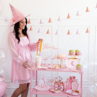 Woman dressed in pink setting up a pink-themed Halloween party table with potion bottle-shaped paper plates and other decorations.