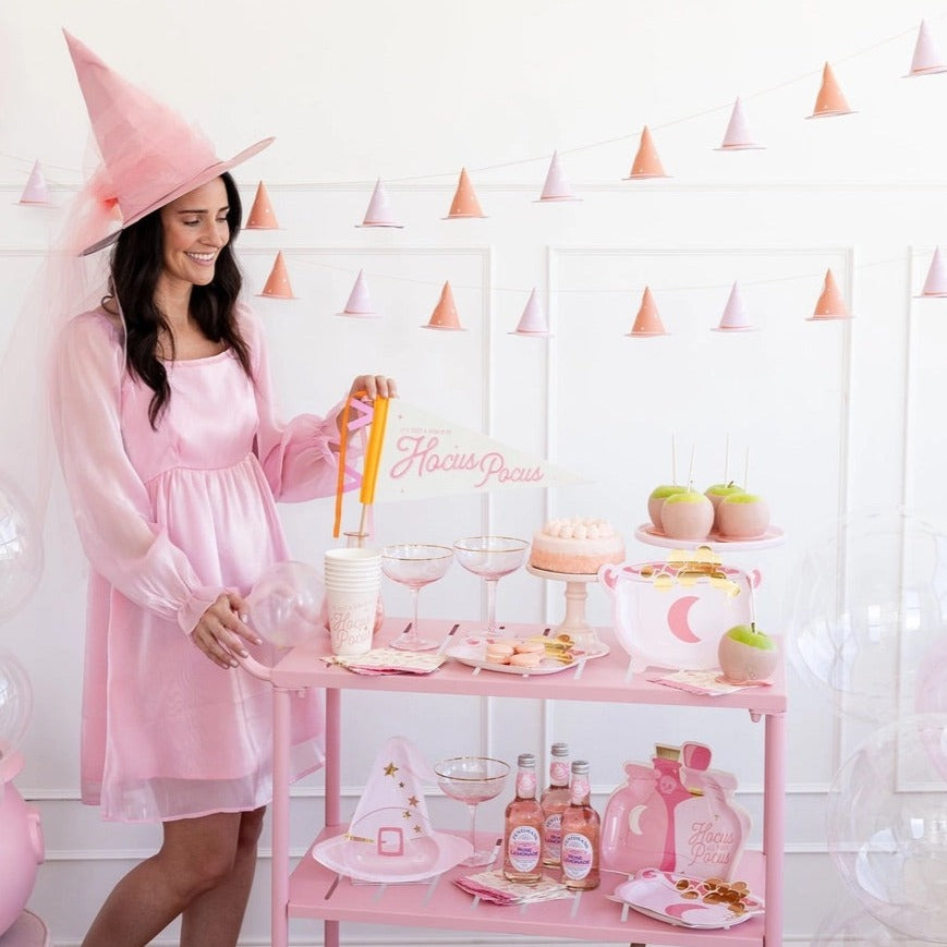 Woman in pink dress setting up a Halloween party table with cauldron and potion bottle-shaped paper plates and pink decorations.