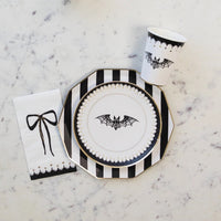 A chic Halloween table setting featuring classy Halloween decorations, including a striped Halloween plate, bat-themed cup, and a white Halloween paper napkin with a black bow design. Perfect for an elegant yet spooky celebration.