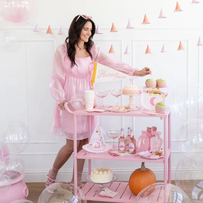 Woman in pink dress with a Halloween party setup featuring cauldron-shaped paper plates, potion bottle-shaped plates, and other pink decor.