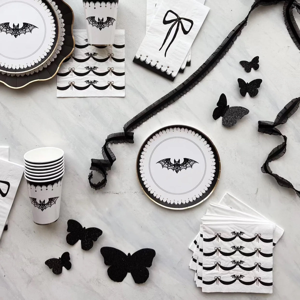 An elegant flat lay showcasing black and white Halloween napkins, plates, and cups with bat and bow motifs. The black butterflies and ribbon add a touch of whimsy to the refined Halloween decor.