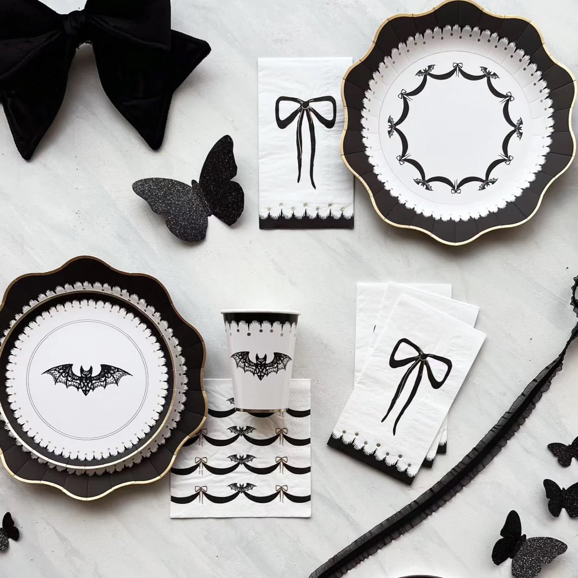 A flat lay of classy Halloween decorations, including black and white Halloween paper napkins with bow designs, bat-themed plates, and cups. The perfect mix of elegance and spooky sophistication for a high-end Halloween party.